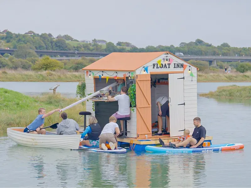 The Float Inn: a small pub with a peach roof and bunting, floating on the water with family on paddleboards and a rowing boat beside it.